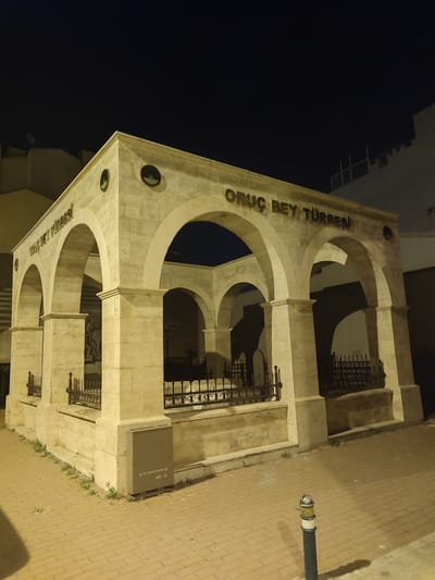 In the city of Osmangazi, Turkey, at night, a stone structure with arched openings is illuminated. The structure has the words "ORUÇ BEY TÜRBESİ" inscribed on its facade in raised, metallic letters. Within the arched bays, black metal fencing encloses areas that appear to be tombs. The ground in front of the structure is paved with brick-like stones. A dark gray electrical box or utility cabinet is visible on the right side of the structure. A reflective bollard stands on the brick pavement in the foreground. The sky above is dark and appears to be clear of clouds. Buildings are visible in the background.
