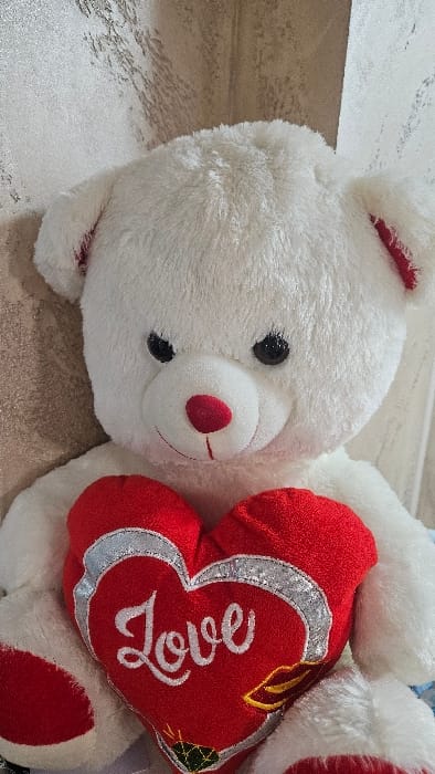 A white plush teddy bear is depicted seated indoors in the city of Iasenovets, Bulgaria. The bear possesses black plastic eyes, a red fabric nose, and a thin black embroidered mouth line. Its inner ears are red. The bear holds a red, heart-shaped plush object against its torso. This heart features a silver perimeter and the word 'Love' embroidered in white script. Below 'Love', golden lips are embroidered to the right and a green gem with golden highlights is embroidered to the left. The background behind the bear consists of a corner formed by two walls. The wall to the left is textured and light beige with an irregular, reflective pattern. The wall to the right is lighter, possibly white, with a faint vertical texture.