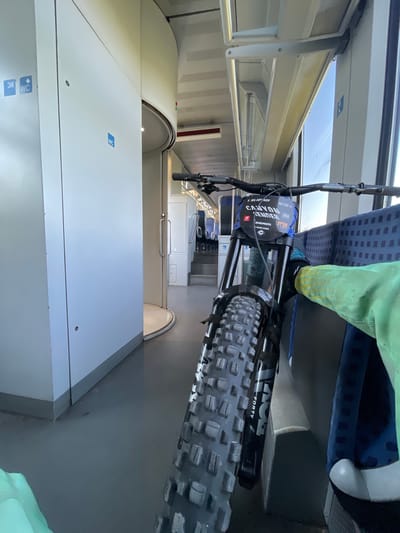 The image shows a mountain bike with its front wheel and handlebars in the foreground, inside what appears to be a train car. The bike is positioned next to a row of seats. The interior of the train is visible, including the windows, ceiling, and a hallway with a curved wall and door. There are blue "WC" signs on the wall to the left. A person's leg wearing a bright green jacket is visible to the right of the bike, resting on a seat. The lighting suggests it is daytime, with natural light coming from the windows. The overall scene depicts a mode of transport for the bicycle.