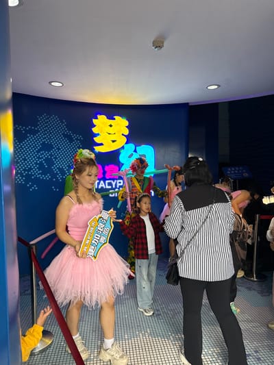 This image depicts an indoor scene within an entertainment or attraction venue located in Shinan District, China. The environment features a curved blue wall adorned with illuminated yellow and blue Chinese characters, notably "梦幻" (Dream/Fantasy), and the English text "TACYP" beneath them, alongside a dotted world map graphic. The ceiling is white, equipped with recessed lighting fixtures and a smoke detector. The floor is covered with light blue tiles arranged in a grid pattern.
In the foreground to the left, a young adult female entertainer stands, dressed in a sleeveless pink top with feathered trim and a voluminous, layered pink tulle skirt, paired with white socks and platform sneakers. Her hair is styled in braids, topped with a colorful floral headpiece. She holds a white and blue sign displaying Chinese characters that translate to "Qingdao, a romantic city, plus love."
To her right, a young girl, wearing a red and black plaid shirt over a white top, light blue wide-leg jeans with butterfly embroidery, and silver sneakers, stands facing slightly right. Behind this child, a person in a vibrant clown costume, featuring a plaid top, striped pants, and a multi-colored afro wig, holds yellow and orange balloon animals.
On the right side of the frame, an adult female is seen from behind, wearing a black and white vertically striped shirt with a black yoke and black pants. She carries a black shoulder bag with a chain strap and observes the scene. In the bottom-left corner, a child's hand in a yellow sleeve is partially visible, reaching up from behind a red barrier rope. The overall scene suggests a family-friendly event with costumed performers interacting with visitors.