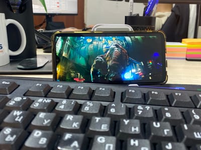 The image shows a close-up view of a smartphone displaying a scene from the Teenage Mutant Ninja Turtles movie, placed horizontally on a black keyboard. The smartphone is supported by a white object at the top. In the background, a white mug with a black handle is visible to the left of the phone. Further back, a computer monitor and a pen holder with blue pens and a pencil are present. To the right of the phone, stacks of colorful sticky notes are arranged. The scene is set on a desk in what appears to be an office environment. There is no indication of the city of YUKHTA, RUSSIA.