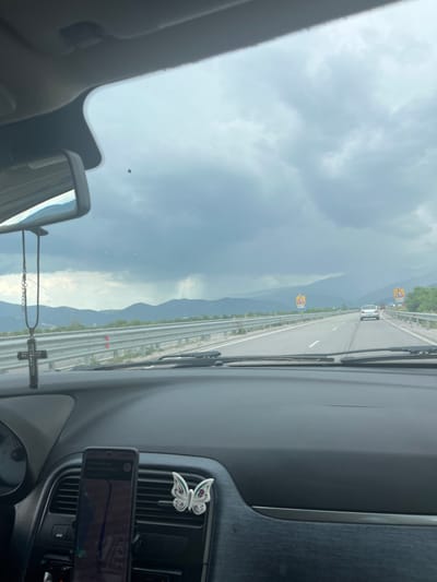 The image is taken from the interior of a car, looking out through the windshield and side mirror. The view is of a highway with a silver car and a truck in the distance. The sky is overcast with dark, stormy clouds. Mountains are visible in the background. There are yellow triangular road signs visible on the right side of the road. Inside the car, a phone is mounted on the dashboard, displaying navigation. A decorative white butterfly and a black cross hanging from the rearview mirror are also visible. The location is the highway near Skravena, Bulgaria.