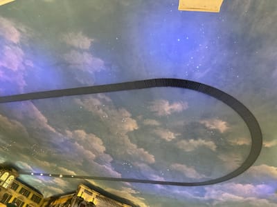 The image shows a ceiling painted with a sky mural, featuring clouds and stars. There is a curved, grey, ribbed structure running across the ceiling. Below this, another similar structure is visible, with lights illuminating it. In the lower part of the image, architectural elements resembling buildings with windows and balconies are depicted, suggesting an indoor space designed to mimic an outdoor scene. The overall impression is of an elaborate interior design, possibly in a themed environment. The specific location in Trafford, United Kingdom, is not discernable from the image itself.
