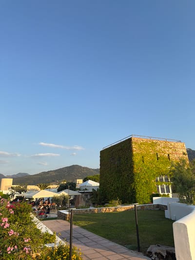 This is a vertical, eye-level, medium-shot photograph taken outdoors in Finale, Italy, during the day. The sky is a clear, bright blue with a few wispy white clouds scattered across the upper portion of the image. In the foreground, a pathway made of light-colored square tiles leads into the scene. To the left of the pathway, a lush garden area is filled with vibrant pink and yellow flowers and green foliage.
Dominating the right side of the image is a square, stone tower covered in dense green ivy. The tower has a flat roof surrounded by a metal railing. Several rectangular windows with white frames are visible on the ivy-covered side. To the left of the tower, the scene opens up to an outdoor seating area under large white umbrellas. Several people are seated at tables, engaged in dining or conversation. In the background, the landscape features rolling green hills and several buildings with light-colored facades and white tents. The overall atmosphere suggests a pleasant, possibly resort-like, setting.