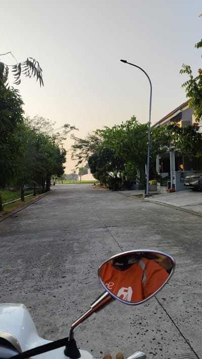 The image captures a street scene in the city of Kebalen, Indonesia, likely during the golden hour due to the soft, warm lighting. A paved road stretches into the distance, lined with lush green trees on both sides. On the right, a modern house with a dark roof and light-colored walls is visible, with a car parked in its driveway. A street lamp curves over the road. In the foreground, the chrome mirror of a white scooter reflects a person wearing an orange jacket with a white logo, indicating they are likely a delivery rider. The sky is a pale, clear blue, transitioning to a warmer hue towards the horizon. The overall impression is one of a quiet, suburban street at either dawn or dusk.
