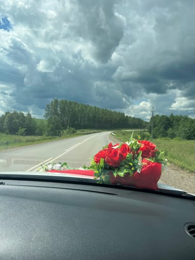 A bouquet of red roses and white flowers, tied with a red ribbon, sits on the dashboard of a car. The car is driving down a paved road lined with green trees and grass. The sky is cloudy and gray. The location is the city of Yunost', Russia.