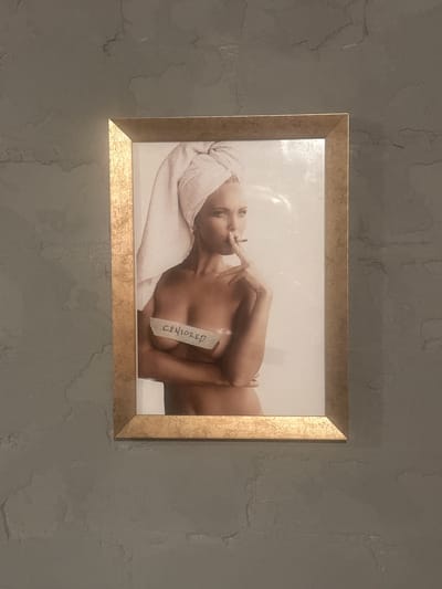 A framed photograph is mounted on a textured gray wall. The photograph features a woman with a towel wrapped around her head, holding a cigarette to her lips. A strip of tape with the word "CENSORED" written on it is placed over her chest. The frame is golden and appears to be made of metal. The wall has a rough, mottled texture with variations in shade. The image and its placement are consistent with decorative artwork in a private residence or establishment in Lapės, Lithuania.