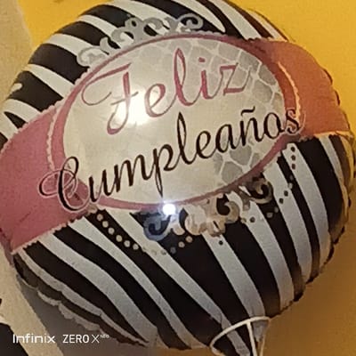 A foil balloon with black and white zebra stripes. A pink banner with a white oval containing "Feliz Cumpleaños" in a swirling script font, the "Feliz" in pink and "Cumpleaños" in black. The background of the oval features a subtle gray, patterned design. The balloon appears slightly deflated. The bottom of the balloon has gathered material tied closed. "Infinix ZERO X Neo" is visible at the lower left corner of the image.