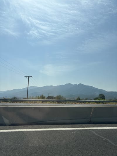 The image captures a scene from the perspective of a moving vehicle on a highway, likely in or near Tresserre, France, given the hazy mountain range in the background. The foreground is dominated by the asphalt of the highway, marked by a white line on the right side. A concrete barrier runs horizontally across the midground, separating the highway from the landscape beyond. Behind the barrier, a metal guardrail is visible. A single utility pole with multiple wires stretches across the left side of the frame, against a backdrop of rolling hills and a distant, silhouetted mountain range that appears to be covered in vegetation. The sky above is a pale blue, sparsely populated with wispy, white clouds that are characteristic of fair weather. The overall impression is one of a rural or semi-rural highway scene, potentially during daylight hours given the bright sky. There are no people or animals visible in the frame, and the focus is on the landscape and infrastructure.