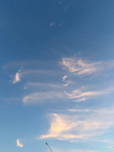 The image shows a clear blue sky with wispy, light-colored clouds. A black lamppost is visible in the lower right corner, with a portion of a dark building or structure in the far lower right corner. The lighting suggests it is either sunrise or sunset. The scene is an outdoor environment, likely a city, given the presence of the lamppost and building. There are no people or animals visible in the image.