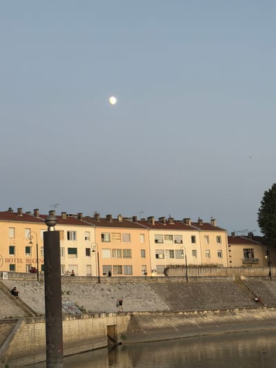 The image depicts a waterfront scene in Arles, France, likely at dusk given the lighting and the presence of the moon in the sky. A row of apartment buildings with red-tiled roofs lines the background. Some of these buildings have balconies, and their facades are painted in pale yellow and beige tones. One building prominently displays a sign reading "HOTEL REGINA."

In the mid-ground, a stone embankment separates the buildings from the water. Several street lamps are visible along the embankment. A large, dark cylindrical object, possibly a metal pipe or a decorative element, stands vertically in the foreground, partially obscuring the buildings. There are steps leading down to the water on the embankment.

Several individuals are present. Two people are seated on the steps on the left side of the embankment. Another person stands on the embankment near the middle, and two more people are walking on the embankment on the right side. In the water, reflections of the buildings and the sky are visible. The sky is a pale, hazy blue, with the moon appearing as a bright, diffused orb. A few distant objects, possibly birds or planes, are faintly visible in the sky.