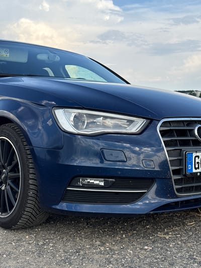 The image shows the front left quarter panel, headlight, grille, and tire of a dark blue Audi A3 sedan parked on a gravel surface. The car's license plate, partially visible, reads "G D B". The background features a cloudy sky and a hint of a grassy embankment in the distance. There are no people or discernible actions occurring in the scene. The car appears to be in a rural or semi-rural setting, possibly on the outskirts of a town like Hahn, Germany, given the provided location.