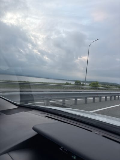 The image depicts a view from the interior of a vehicle, looking out towards a highway and a body of water under a cloudy sky. The dashboard of the car is visible in the foreground. A metal guardrail runs along the side of the highway, separating it from a grassy area and a body of water. In the distance, there are trees and hills. A lamppost is visible on the right side of the frame, extending into the sky. The overall impression is one of travel on a highway near a large body of water. The location is identified as De-Friz, Russia.
