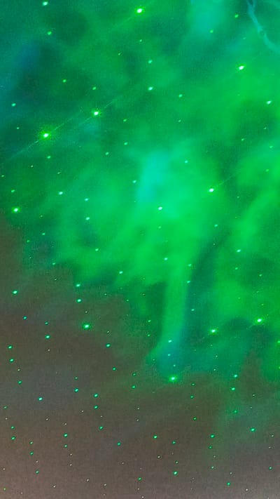 The image depicts a dark background illuminated by scattered green lights, resembling a starry sky or a galaxy projection. There are no people or identifiable objects present. The lighting creates a hazy, ethereal effect with areas of brighter green glow and dimmer, more diffused light. It's impossible to determine the specific location in Zvenigorod, Russia, from this abstract imagery.