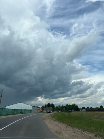 A photograph taken in Голацкі сельскі Савет, Belarus, depicts a roadside scene under a sky dominated by dramatic, dark storm clouds. A two-lane asphalt road curves gently, marked by a solid white line separating the lanes. Adjacent to the road is a grassy verge transitioning into a field. On the left side of the road, structures, including a long, low building with a light-colored corrugated roof and green fencing, are visible. Power lines are present. The sky features a mix of blue patches and dense, gray clouds.