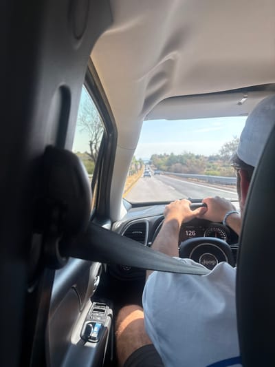 Inside a car, a driver in a light-colored shirt and dark shorts grips the steering wheel, with a digital display reading "126" visible on the dashboard. A prominent seatbelt bisects the view. Outside the window, a highway stretches with greenery and distant vehicles. The car's interior, including the headliner, is visible, along with controls and vents. The focus is on the driver's actions and the road ahead.