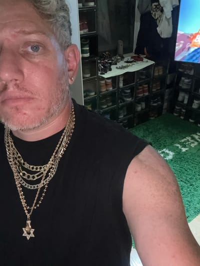 An adult male is depicted in a close-up, selfie-style photograph, showing his upper torso and face. He possesses light-colored eyes and prominent graying, light-colored hair, with a short beard or stubble on his face. He is wearing a plain black short-sleeved or sleeveless top. Around his neck, he wears a layered collection of multiple gold-colored chains of varying styles, with the longest chain featuring a pendant shaped like a hand holding a Star of David, encrusted with clear, small, reflective stones. A small, silver-colored stud earring is visible in his left ear. The background reveals an interior room, likely a walk-in closet or dressing area, characterized by vertical stacks of clear storage boxes containing numerous pairs of shoes/sneakers. A flat surface in the mid-ground holds various small objects. To the right, a television screen displays vibrant, colorful digital content. The floor is covered with a green, shaggy rug featuring white markings, reminiscent of a sports field. The scene is located in Alfey Menashe, Palestinian Territory.