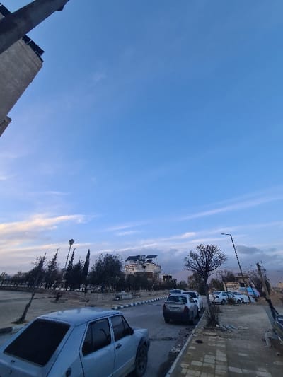 A street scene under a vast blue sky with light clouds. Several cars are parked along the road, with a light-colored vehicle prominently featured in the foreground. A row of trees lines the street, and buildings are visible in the background, nestled beneath the hills. A lamppost stands along the roadside. The image is rotated, with the sky taking up a large portion of the frame.