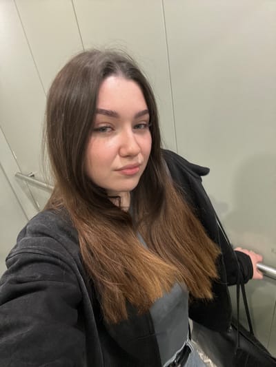 A young woman with brown hair is looking at the camera. She is wearing a black jacket and appears to be holding onto a metal bar. The background is a plain, light-colored wall. It is unclear if the location is Osentsy, Russia, based on the image alone. No other specific objects, actions, or interactions are directly observable.