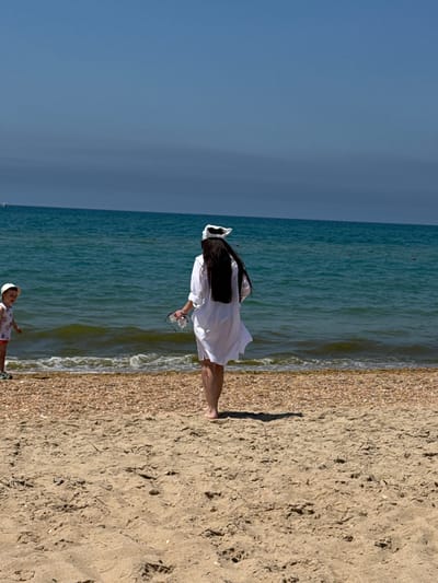An image taken on a sunny day shows a beach in Vityazevo, Russia. In the foreground, the sandy beach transitions to a pebble-strewn shoreline meeting the turquoise waters of the sea. A young child, wearing a white hat and a patterned top with red shorts, stands near the water's edge, looking towards the right. Further back, a woman in a white cover-up and a white headscarf stands with her back to the camera, holding sunglasses and a clear bottle. The sea is calm, with small waves breaking on the shore. The sky is a clear, pale blue.