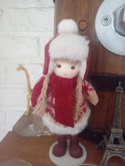 A close-up, slightly angled shot captures a small fabric doll dressed in winter attire, standing on a wooden shelf. The doll has a light-colored face with small black eyes and a stitched red smile. It wears a red hat with a white fur trim and a matching white pom-pom on top. A red scarf is draped around its neck, and it's dressed in a red coat with white snowflake patterns and white fur trim. The doll's hair is made of light brown yarn braided into two pigtails.
Behind the doll, to the left, is a blurred, light-colored brick wall. To the far left and slightly out of focus, a small, yellow toy giraffe stands on a clear glass object. To the right of the doll, a dark wooden surface is visible, possibly part of a larger object or furniture. A small, metallic Eiffel Tower replica stands on this surface. The overall lighting is soft, suggesting an indoor setting.
The image's metadata indicates it was taken on December 24, 2021, at 11:32 AM, with a Samsung SM-A525F camera. The GPS coordinates provided place the location in Terebovlia, Ternopil Oblast, Ukraine.