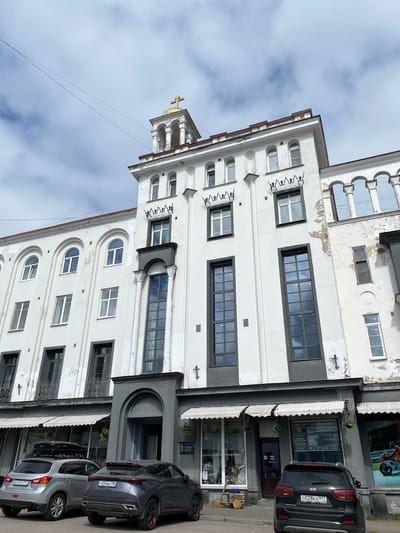 The image shows a multi-story white building with arched and rectangular windows, located in Sortavala, Russia. The building has architectural details such as pilasters, decorative elements above some windows, and a small balcony. At the top of the building, there is a small cupola topped with a golden cross, indicating a religious affiliation or architectural style. Several cars are parked in front of the building, and there are awnings and shop windows on the ground floor. The sky is cloudy.