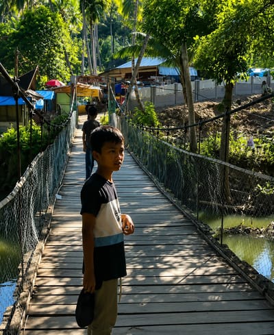 A young boy stands on a wooden suspension bridge in Silaiyajae, Indonesia, looking to his right. He is wearing a black t-shirt with white and light blue horizontal stripes on the chest and khaki pants. He holds a black hat in his left hand. The bridge is made of weathered wooden planks and has wire mesh railings. Sunlight casts dappled shadows across the bridge's surface. In the background, more children are visible walking on the bridge, and a lush, green tropical landscape with palm trees and vegetation borders the scene. Small buildings with blue and red roofs are visible on the far bank of the river below the bridge. The water in the river is murky green.