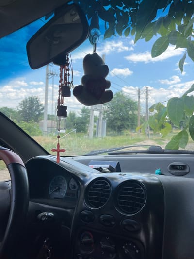 The image shows the interior of a car, viewed from the passenger seat looking towards the dashboard and windshield. Outside, the sky is blue with scattered white clouds, and green foliage is visible, suggesting a daytime setting with fair weather. The scene appears to be set in a rural or suburban area, with trees and utility poles visible through the windshield, consistent with the provided location context of Chernove, Ukraine.
Inside the car, several objects are visible. A rearview mirror hangs from the windshield, with a teddy bear ornament attached above it. Below the mirror, prayer beads with a small glass bottle and a red cross charm are hanging. The dashboard features air vents, a digital clock displaying "10:01", and a portion of the instrument cluster, including a speedometer. The steering wheel is partially visible in the foreground. No people are present in the image. The overall impression is one of a parked vehicle on a sunny day, with personal decorative items adorning the interior.