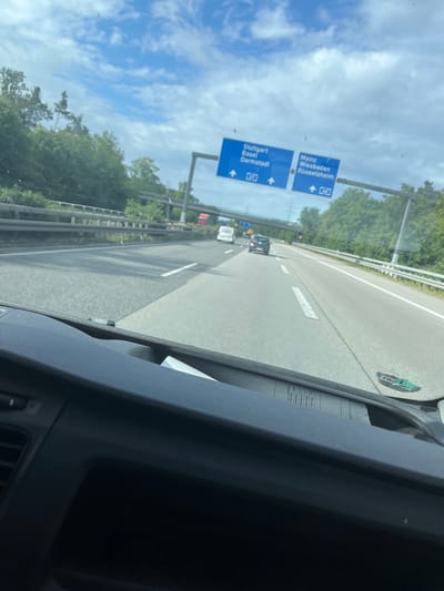 The image captures a first-person perspective from inside a vehicle, looking out onto a multi-lane highway (Autobahn) under a bright, partly cloudy blue sky during the daytime. The scene is set in Germany, near Rüsselsheim am Main, as indicated by the road signs.

The primary subjects are the highway stretching into the distance, surrounded by green trees, and two blue overhead road signs. The lower sign directs traffic to "Stuttgart," "Basel," and "Darmstadt" via the "A 67" highway, with an arrow indicating the route continues straight. Above it, another sign points towards "Mainz," "Wiesbaden," and "Rüsselsheim" on the "A 60" highway, suggesting a turn-off or split. In the distance, several vehicles are visible on the highway.

The activity is driving on an Autobahn. The weather is clear and sunny, with scattered white clouds. Notable details include the dark gray interior of the vehicle in the foreground, specifically the dashboard and part of the A-pillar on the right. A document or piece of paper is tucked into a holder on the dashboard. Visible text from this document includes "Verhundert," "Geibel Transp," "Bozhidar Nik," "Vorschuss M," and "Weterstad." The highway itself has clear white lane markings.