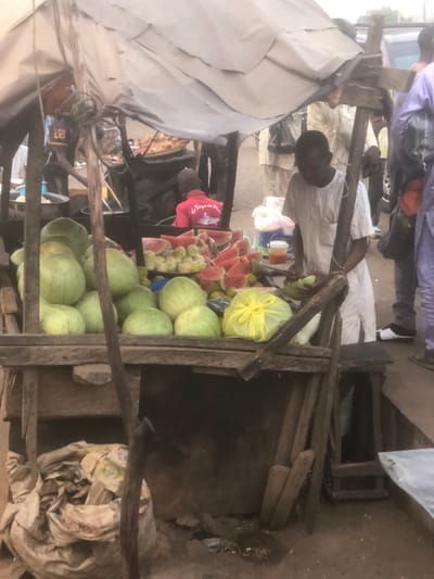 An outdoor market scene in Tukur Tukur, Nigeria, depicts a fruit stall with a makeshift tarp canopy. A dark-skinned male in a white tunic stands behind the wooden stall, actively preparing or cutting produce, possibly peeling a green item. The stall displays numerous whole, light-green watermelons, piles of cut red watermelon slices, and mounds of round yellow fruit. A yellow plastic bag is present on the stall surface. Underneath the stall, several sacks are visible on the unpaved, earthen ground. In the immediate background, another individual wearing a red shirt with white lettering is seated behind a separate food stall, where fried items are visible in a metal pan. Further back, several indistinct figures, some in light-colored traditional garments, are present, indicating general market activity. A vehicle is partially visible in the far background on the right.