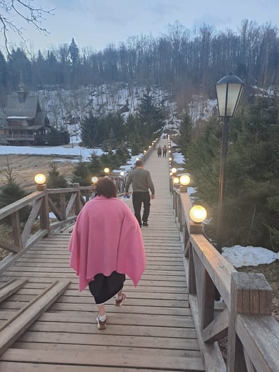 The image shows a downward-sloping wooden pedestrian bridge in what appears to be Vzglyadnevo, Russia. Several people are walking on the bridge. One individual, closest to the camera, is wearing a pink garment. Another person, farther down the bridge, is wearing green and black clothing. The bridge is lined with evenly-spaced, spherical lamps. The surrounding environment consists of trees, some bare and some evergreen, as well as areas with snow cover. A building is visible at the base of the bridge. A tall street lamp stands at the bridge's highest point.