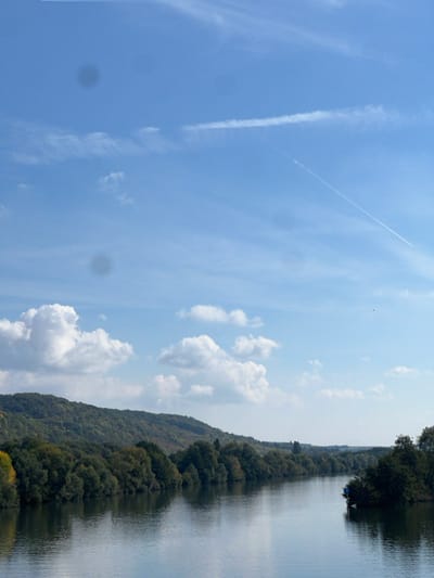 The image displays a wide landscape scene of the Seine River near Vernon, France, observed under a bright blue sky. The sky contains scattered white cumulus clouds, wispy cirrus clouds, and two distinct aircraft contrails: one long, thin, and diagonal across the upper right, and another broader, fading one in the upper left. Several dark, circular artifacts, indicative of lens flares or sensor dust, are visible against the sky.
The background consists of rolling hills covered in dense, dark green deciduous forest, with some trees showing hints of yellowing foliage, suggesting an autumn season. The wide, calm river occupies the mid-ground and foreground, reflecting the blue sky and the trees on its banks. Both river banks are extensively lined with mature trees. On the right side of the river, positioned near the bank amidst the trees, a small, dark-colored object, possibly a boat or a fixed structure, is visible with a blue covering or tarp on its upper surface. Small, dark specks, consistent with distant birds, are sparsely distributed in the sky. There are no discernible human figures, actions, or interactions within the frame.