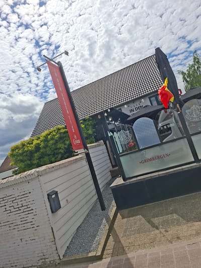 The image depicts the exterior of a building with a tiled roof, located in Opglabbeek, Belgium. The building appears to be a establishment with a terrace area, evidenced by outdoor seating, glass enclosures with branding, and an umbrella. A red banner with text and a logo hangs from a pole in the foreground. A Belgian flag is visible on the right side of the building, attached to an umbrella. The surrounding environment includes a white brick wall with a mailbox, gravel ground, and paved areas. The sky is filled with scattered clouds. There are no people visible in the image.