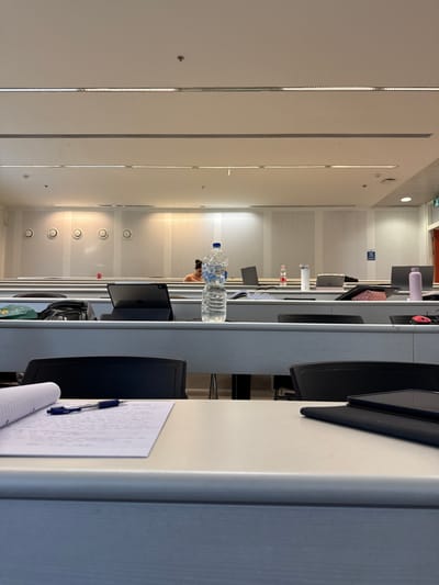 The image shows a classroom at Kfar Monash, Israel.  A single, partially obscured person is sitting at a desk, facing away from the camera. The person is looking at a computer screen. A plastic water bottle is sitting on the desk, and a notebook is lying on the desk in front of the camera. There are two chairs in the foreground. The walls are white and there is a ceiling with multiple fluorescent lights. The room is organized in rows of desks.  A red folder is visible in the background, near the person. The room appears to be empty except for the one person. The coordinates (latitude: 32.342, longitude: 34.911) place the location in Kfar Monash, Israel.