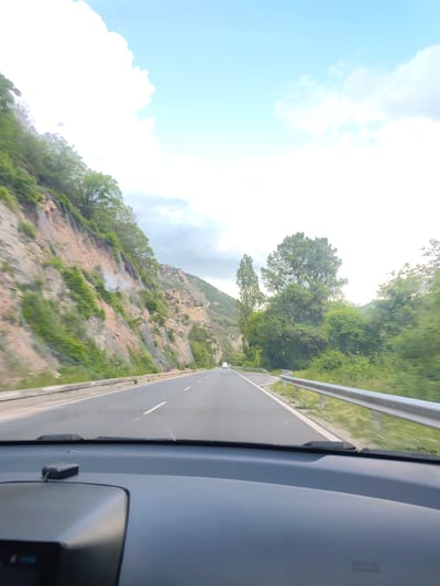 The image depicts a view from inside a vehicle traveling on a highway. The vehicle is moving through a mountainous region near Gospodintsi, Bulgaria. The road is asphalt with white lane markings and a metal guardrail on the right side. To the left, a steep rock face covered with vegetation rises. On the right, trees and bushes line the roadside. A single vehicle is visible in the distance on the road ahead. The sky is visible above the mountains, showing clouds and blue patches. The car's dashboard is visible in the foreground.