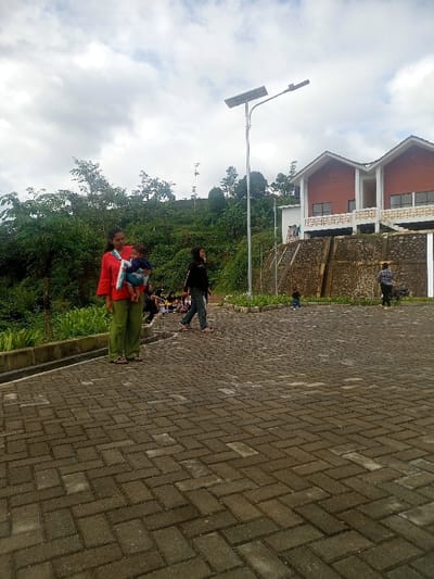 The image displays an outdoor scene in Pangalengan, Indonesia, under an overcast sky. The foreground and middle ground are dominated by a paved area constructed with dark grey interlocking pavers laid in a herringbone pattern.
In the left foreground, a woman wearing a red shirt and green pants stands, holding a baby securely in a blue carrier. The baby faces away from the camera. To her right and slightly behind, another woman in a dark top and jeans walks towards the right side of the frame, appearing to be talking or smiling. Further to the left, a group of individuals, possibly children or young adults, are seated on the ground near a low retaining wall, mostly obscured by the standing woman.
A prominent feature in the mid-ground is a tall, grey street light pole equipped with a solar panel on top, indicating a solar-powered lighting system. To the right of the pole, a multi-story building is visible, featuring two distinct gabled roofs, red upper walls, and white balconies or structures below. This building is situated on higher ground, separated from the paved area by a stone retaining wall.
The background is characterized by lush, green hills densely covered with trees and vegetation, typical of a mountainous or hilly region. Two more figures are visible near the building on the right: one is a small child in a blue jacket, and another person is partially obscured further to the right. The scene depicts a gathering of people in what appears to be a public or recreational area, enjoying the outdoors during daylight hours on a cloudy day. There is no discernible text visible in the image.