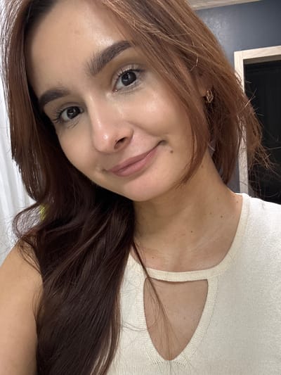 A close-up, selfie-style photo captures a young woman with auburn hair and fair skin. She is looking at the camera with a slight smile, her head tilted to her left. Her makeup includes mascara and a matte, nude lipstick. She is wearing a cream-colored knit top with a keyhole cutout at the neckline. The background is slightly blurred, showing a dark wall with a doorframe and a white curtain. There is no indication of the location being in Kuyuki, Russia.