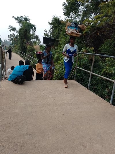 A group of people traverse a concrete walkway bordered by metal railings and lush greenery. Several women carry items on their heads, including baskets, buckets, and a large bundle. Others sit alongside the walkway, one partially obscured by a blue cloth and a black bag. A solitary figure in dark clothing walks away in the distance. The lighting suggests a cloudy or overcast day.