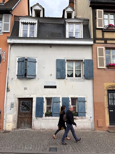 The image shows a narrow street in Colmar, France, with traditional Alsatian architecture. The facade of a building is prominent, featuring a light-colored plaster wall with blue shutters on its windows. A sign above one window reads "POISSONNERIE," indicating a fish shop. Two people, a woman and a man, are walking from left to right on a cobblestone sidewalk. The man wears a dark sweater and blue jeans, carrying a backpack. The woman is dressed in black. The street is lined with multi-story buildings, one with an orange facade and another with a tan, timber-framed facade. The sky is overcast.