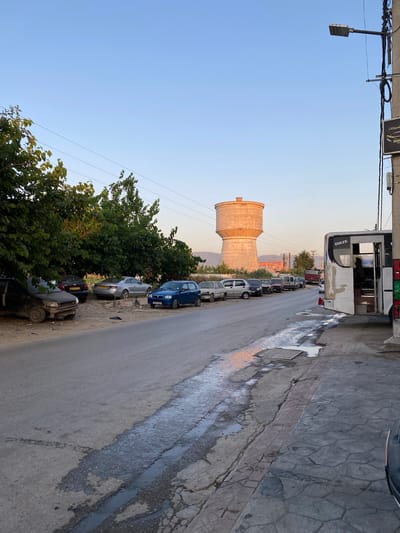 The image captures a street scene in Sidi Moussa, Algeria, during what appears to be late afternoon or early evening, characterized by warm, soft lighting. A prominent, large cylindrical water tower stands in the mid-ground on the left, illuminated by the low-angled sun.

Along the right side of the paved road, numerous vehicles are parked in a line, including a blue hatchback, a silver sedan, a black sedan, and other cars of various models. A white and grey bus, partially visible on the far right, also appears to be parked, with "ISUZU" clearly readable on its side.

Overhead, power lines and a street light fixture are visible against a clear, bright blue sky. A sign attached to the overhead structure displays Arabic text, including "للسيارات" (for cars), and the English word "DINAMO," suggesting a business related to automobiles.

The road surface exhibits wear and tear, with cracks and patched areas. Several puddles of water are scattered across both the road and the adjacent textured concrete sidewalk, some reflecting the warm light. Lush green trees line the left side of the road, partially obscuring some of the parked cars. In the distance, faint mountains or hills are visible under the clear sky. No people are visible in the scene.