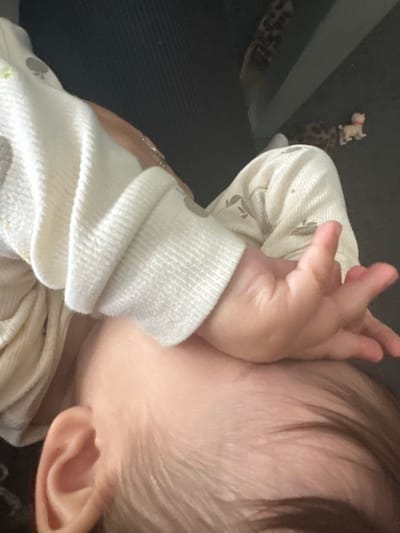 A close-up image shows a baby lying on their side. The baby's right ear is visible, as well as the back of their head and some hair. The baby's right hand is near their face. The baby is wearing a light-colored, long-sleeved garment. In the background, out of focus, are some items, including what appears to be a small toy dog and a patterned surface.
The image was not taken in the city of Macclesfield, United Kingdom, as there is no indication of location.