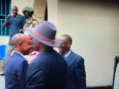 The image depicts a group of four men standing in front of a white wall in a building in Keshero, Democratic Republic of the Congo. One of the men is wearing a blue hat and a dark suit jacket. Another man is wearing a light blue shirt and a dark suit jacket. The two men are looking at each other and smiling. There are two additional men in the background. The man on the left is wearing a dark blue shirt and the man on the right is wearing a light blue shirt. A statue of a soldier wearing a helmet and camouflage is positioned in the background. A light blue plastic chair is positioned in the background. The scene is shot in a close-up perspective.