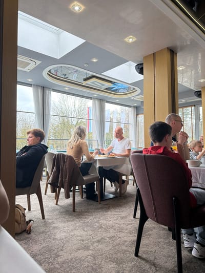 The image shows an interior scene, likely a restaurant or dining area, with several individuals seated around tables. The location is possibly Vidin, Bulgaria, as suggested by the flags visible through the windows, but this is not definitively confirmed. There are multiple people in the image: A person in a brown jacket is seated at a table, appearing to be deep in thought or looking down. Adjacent to them, a woman with blonde hair is seated at the table, possibly interacting with someone. Across the table, a man in a white shirt appears to be handling food. In the background, there's another table where a person in a red shirt is seated, and next to them, a bald man is engaged in conversation with a young boy. The environment is well-lit with large windows providing a view of the outside, where trees are visible. The decor of the dining area includes an artistic design on the ceiling, and the room is furnished with modern chairs and tables. A purse is on the floor. The overall impression is a gathering of people, possibly a family or group of friends, enjoying a meal together.