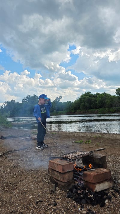 A young boy wearing a blue baseball cap and jacket stands on a gravelly riverbank, holding a stick and looking towards the water. In the foreground, a makeshift grill made of bricks is set up with a fire burning underneath, cooking what appears to be meat on a grill rack. The river is wide and calm, with a dense green forest lining the opposite bank. The sky is partially cloudy with dramatic cumulus clouds. The location is Дарьино, Russia.