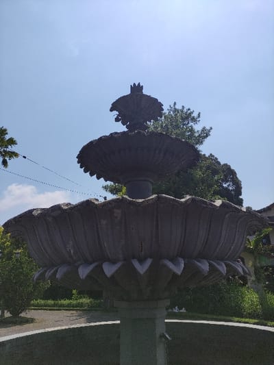 The image depicts a two-tiered, dark gray stone or concrete fountain located in Staufenberg, Germany, under a clear blue sky. The fountain's main visible tier is broad and scalloped, with a pattern of V-shaped white accents along its lower edge. Above this, a narrower, similarly fluted tier supports a crown-like or stylized finial at its apex. The fountain's central column extends downwards into a partially visible circular basin, with a small metallic spout discernible on its lower section. Surrounding the fountain are various green trees and bushes, indicating a park-like or landscaped setting. A paved pathway curves behind the fountain's base. In the upper left quadrant, two parallel dark wires with small white insulators traverse the sky, and a fragment of a lamppost or similar structure is visible. No individuals are present, and no overt actions are occurring.