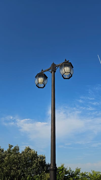 The image depicts a lamp post with two globes against a bright blue sky. The lamp post is black and has an ornate design. The globes are also black with glass panels that allow the light inside to be seen. The light inside the globes appears to be a warm yellow. There are some wispy white clouds scattered across the sky. On the right side of the image, there are green trees. In the upper part of the image, a white contrail is visible in the sky, suggesting an airplane has passed overhead. The image appears to be taken during the daytime, likely in the late afternoon due to the warm light reflecting on the lamp post. The overall mood of the image is serene and peaceful. The location is Cheonan-si, South Korea. There is no visible text in the image.