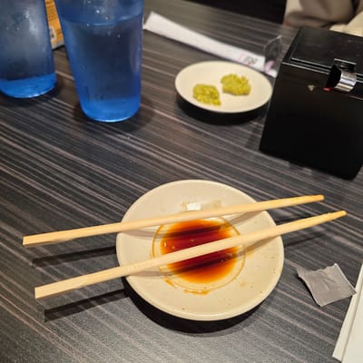A close-up, eye-level shot shows a dark wood grain table with two blue glasses of water and a black napkin holder on the upper right. In the center of the frame, a light-colored plate holds a small amount of soy sauce and two wooden chopsticks resting on top, partially submerged in the sauce. To the left of the plate, two small mounds of green paste are on a smaller white dish. A white paper wrapper lies near the bottom right of the frame. The scene appears to be in a restaurant in Beaverton, Oregon.