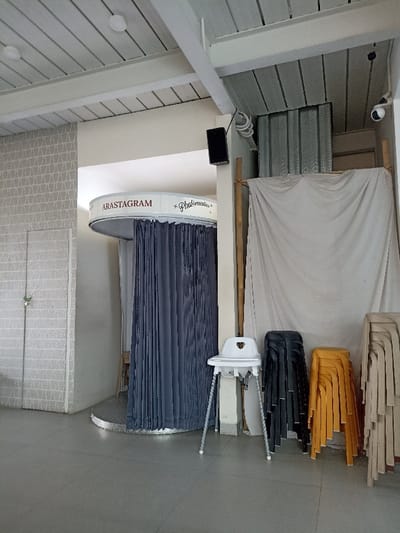 The image shows an indoor photo studio with a sign that reads "ARASTAGRAM - Photomatics." The studio is located in Purwokerto, Indonesia (latitude: -7.42, longitude: 109.245). The studio has gray tiled floors, white walls with a textured pattern, and a white ceiling with wooden beams. There is a black curtain in a photo booth behind a circular white platform. To the right of the photo booth is a white sheet hanging from the ceiling. There are three chairs in front of the sheet. One chair is white, one is black, and one is yellow. The chairs are in front of a white wall with a textured pattern. There is a speaker on the wall to the left of the sheet. There is a white door to the left of the photo booth. The studio appears to be empty and unused.