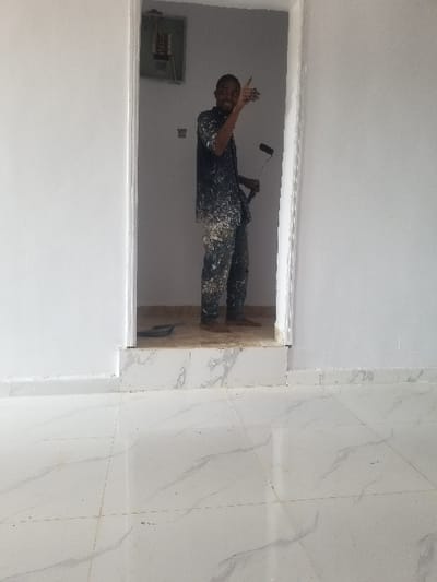 An indoor scene in Edu Town, Nigeria, displays a male individual standing barefoot within an open doorway. He wears a dark short-sleeved shirt and trousers, both heavily splattered with white paint. He holds a black-handled paint roller in his right hand, oriented downwards. His left arm is raised with the index finger extended upwards and to his left, and he is smiling towards the viewer. An electrical service panel is mounted on the white wall visible through the doorway, positioned to the man's upper left. A dark, shallow paint tray rests on the floor just inside the doorway, near his feet. The walls in the foreground and within the doorway are uniformly painted white. The foreground floor is covered with large, light-colored ceramic tiles featuring subtle grey veining, laid in a grid pattern. A matching tiled baseboard runs along the bottom of the foreground wall. The doorway itself has a slightly elevated, tiled threshold.