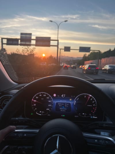The image presents a first-person perspective from inside a Mercedes-Benz vehicle, actively traveling on a multi-lane highway within the city of Amadora, Portugal, at sunset, specifically at 19:51 (7:51 PM). The driver's left hand is visible on the steering wheel, which features the Mercedes-Benz emblem. The digital instrument cluster displays a current speed of 41 km/h. Additional information on the display includes "Após arranque" (After start), indicating a journey consumption of 10.4 l/100km, an average speed of 27 km/h, 23 km driven, and a total driving time of 52 minutes. It also shows "0.0 kWh/100km" and "0% elétrica" (0% electric), with an external temperature of 22.0 °C.

Through the windshield, a low-lying, bright orange sun is visible on the left side of the horizon, casting a warm glow. The highway ahead shows moderate traffic, with numerous vehicles and illuminated brake lights stretching into the distance. Two overhead gantries, likely supporting road signs, span the highway. One gantry features a prominent streetlight with two lamps. The roadside is lined with trees and vegetation, and a distant urban landscape or hills are discernible on the right horizon. The windshield itself exhibits visible dust and streaks.